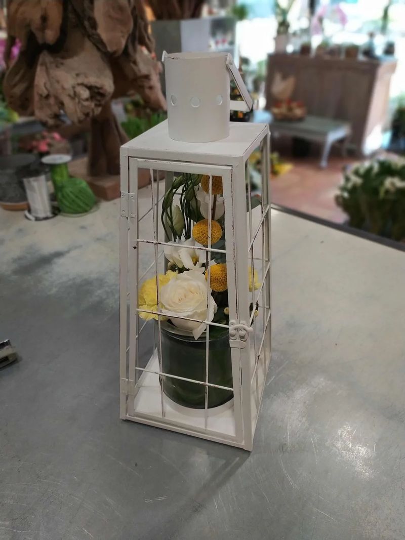 Lanterne de métal blanc garnie de fleurs jaunes et blanches, posée sur le comptoir de zinc de l'atelier, l'intérieur de la boutique en arrière-plan